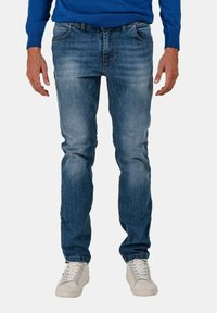 Jeans in denim blu con una vestibilità slim, caratterizzati da un lavaggio sbiadito e cinque tasche. Abbinati a sneakers bianche, con le mani appoggiate sui fianchi.