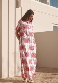 Maxi-jurk met korte mouwen, met een rood palmbomenprint op witte stof. Heeft een losse pasvorm en zakken, gecombineerd met witte sandalen.