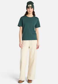 T-shirt à manches courtes turquoise associé à un pantalon large crème, porté avec un bonnet vert orné d'un écusson montagne, le tout dans un style décontracté.