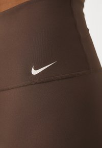Hnědé sportovní legíny z pružné látky, s bílým logem Nike ve tvaru háčka a plochým prošitím podél švů.