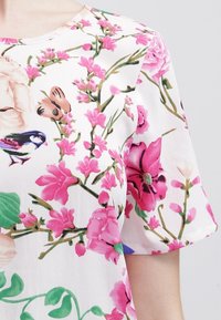 Kwiatowa bluzka z krótkim rękawem z białym tłem, różowymi kwiatami i zielonymi liśćmi, z teksturowanej tkaniny i okrągłym dekoltem.