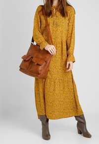 Abito lungo a fiori giallo senape con maniche lunghe, abbinato a una borsa a tracolla in pelle marrone e stivaletti con tacco marroni.