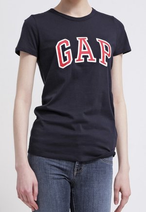 Person wearing a fitted black t-shirt with large white and red "GAP" letters and blue jeans, standing against a plain background.
