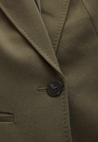 Olivově zelený blazer s texturovaným herringbone vzorem, který má černý knoflík a podrobné šití podél límce a kapsy.