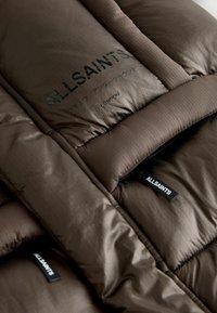Brun vadderad jacka med en jämn yta, detaljerad sömnad och märkta taggar som visar "ALLSAINTS" och "LONDON."