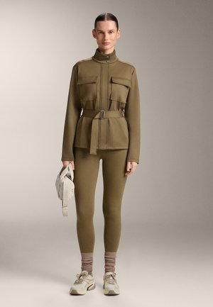 Femeie în picioare purtând o jachetă verde măsliniu cu curea și colanți asortați, ținând o geantă bej, cu adidași bej și șosete maro.