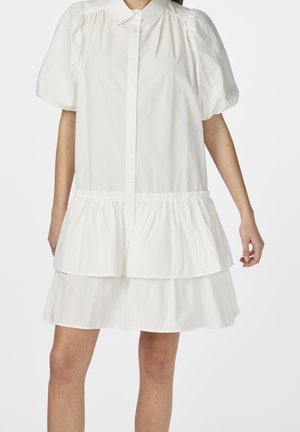 Robe courte blanche avec manches ballons, boutonnée sur le devant, taille froncée et jupe à volants, portée par une personne debout.