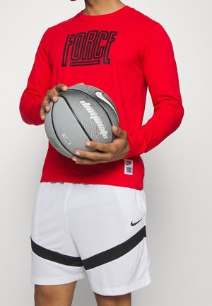 Osoba ubrana w czerwoną, długorękawową koszulkę z napisem "FORCE" oraz białe spodenki do koszykówki marki Nike, trzyma szarą i czarną piłkę do koszykówki Puma.