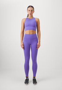 Violetti urheilu crop-top, jossa on korkea kaulus, yhdistettynä samalla värillä varustettuihin korkeavyötäröisiin leggings-housuihin. Materiaali on joustavaa ja sileää.