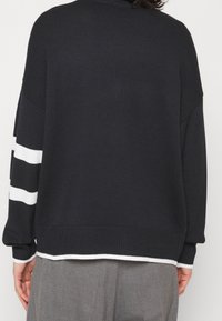 Person wearing a black knit sweater with white stripes on the left sleeve and white trim at the cuffs and hem, paired with gray pants.