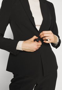 Blazer nero sartoriale con un singolo bottone frontale, revers a punta e pince frontali, indossato sopra un top chiaro a coste.
