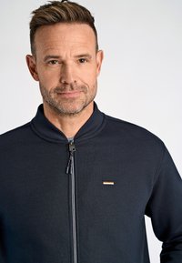 Giacca zip-up di un blu navy scuro con colletto alto, presenta un piccolo dettaglio dorato sul petto e un tessuto texturizzato. Hardware della zip visibile.