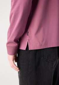 Camisa de manga longa, com decote redondo em algodão rosa claro, com uma fenda lateral e etiqueta de marca na bainha, combinada com calças pretas texturizadas.