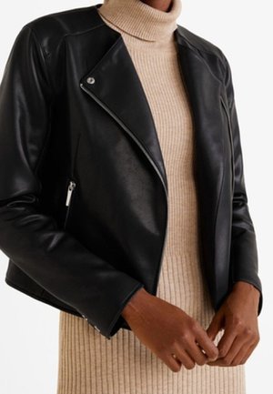 Black leather jacket worn over beige ribbed turtleneck dress, with visible silver zipper and snap button details, hands clasped at waist.