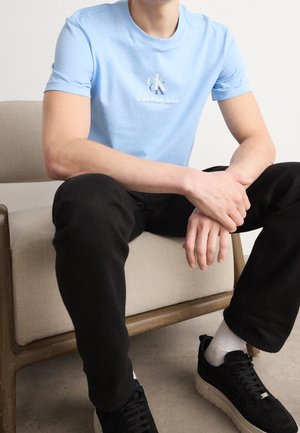 Osoba siedząca na beżowym krześle, ubrana w jasnoniebieską koszulkę Calvin Klein Jeans, czarne spodnie, białe skarpetki i czarne trampki.