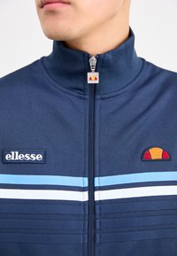 Zbliżenie osoby w granatowej kurtce z suwakem, z jasnoniebieskimi i białymi paskami oraz logo ellesse na klatce piersiowej i suwaku.
