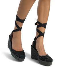 Sandales à talons en wedge noires avec texture tissée, bout fermé et brides croisées à la cheville. Présente des accents décoratifs sur le dessus et une semelle texturée.