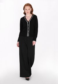 Cardigan negru cu decolteu în V, cu un contur alb în contrast și închidere cu nasturi, asortat cu pantaloni negri evazați și tocuri negre.