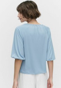 Lys blå kortærmet bluse med rund hals, puffede ærmer og knapdetalje på ryggen. Glat stoftekstur.