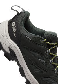 Scarpa da trekking verde scuro con tessuto testurizzato, colletto imbottito, dettagli in rete e suola in gomma grigia. Presenta lacci gialli e branding sul lato.