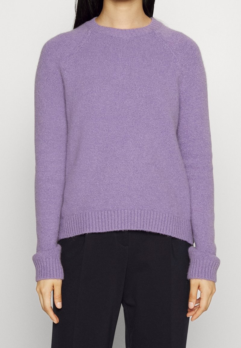 Pull en tricot violet avec un col rond, des poignets et un ourlet côtelés, fabriqué en matériau doux avec une finition légèrement texturée. Associé à un pantalon noir.