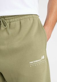 Olijfgroene sweatpants met een elastische tailleband, zijzakken en een witte "new balance" logo gedrukt op de onderkant van het linkerbeen.