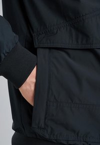 Sort nylonjakke med ribbet manchet, som har en hånd i en sidelomme. Glat tekstur med synlige syninger langs sømmene.
