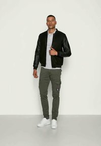 Chaqueta bomber negra con mangas de cuero, camiseta polo gris, pantalones cargo verde oliva con parche y zapatillas blancas. Diseño simple y funcional.