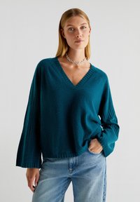 Pulover supradimensionat de culoare teal, cu decolteu în V, țesătură texturată și mâneci lungi evazate. Asortat cu blugi deschiși la culoare, cu talie înaltă.