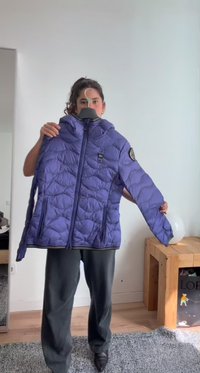 Personne debout à l'intérieur tenant une veste matelassée violette sur un cintre, avec une table en bois et un sac de courses en arrière-plan.