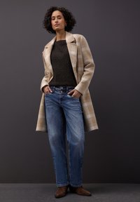 Beżowy wełniany płaszcz w kratę na ciemnobrązowym swetrze z dzianiny, zestawiony z luźnymi niebieskimi dżinsami i brązowymi botkami za kostkę. Prosty design i faktury.