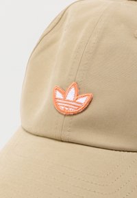 Μπεζ καπέλο μπέιζμπολ με κεντημένο πορτοκαλί και λευκό λογότυπο τριφυλλιού κεντραρισμένο πάνω από την κυρτή γείσο.