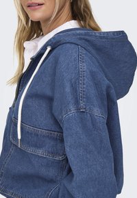 Džínová bunda s kapucí v středně modré barvě s přední kapsou, bílými šňůrkami a viditelnými švy. Volný střih s jemnou texturou.