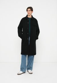 Cappotto nero con colletto, chiusura frontale con bottoni e taschino sul petto. Indossato sopra una camicia verde e jeans azzurri chiari con sneakers bianche.