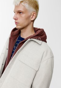 Lichtgrijze wollen jas met een kraag, voorzien van twee voorzakken en een rits, gedragen over een bordeauxrode hoodie en een gestreept marineblauw shirt.