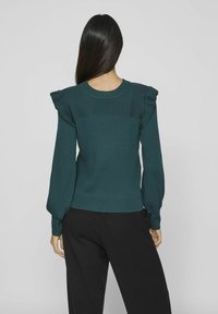 Femme aux longs cheveux foncés portant un pull côtelé bleu sarcelle avec des épaules volantées et un pantalon noir, debout devant un fond clair uni.