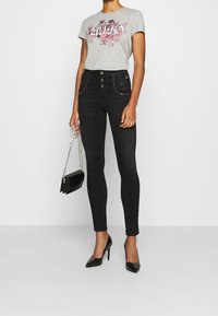 Camiseta gris con estampado floral, vaqueros negros de talle alto con cinco botones, bolsillos adornados y un bolso de mano negro con cadena. Tacones altos.