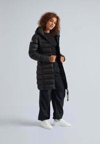 Giacca a vento nera con cappuccio, dotata di una finitura leggermente lucida, chiusura con zip e dettaglio con etichetta. Indossata sopra un top nero e pantaloni neri larghi.