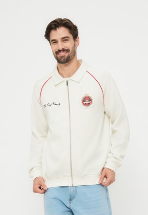 Άνδρας που χαμογελά φορώντας λευκό μπουφάν με φερμουάρ, κόκκινη διακόσμηση, κεντημένο έμβλημα και ανοιχτόχρωμα τζιν, μπροστά σε απλό φόντο.