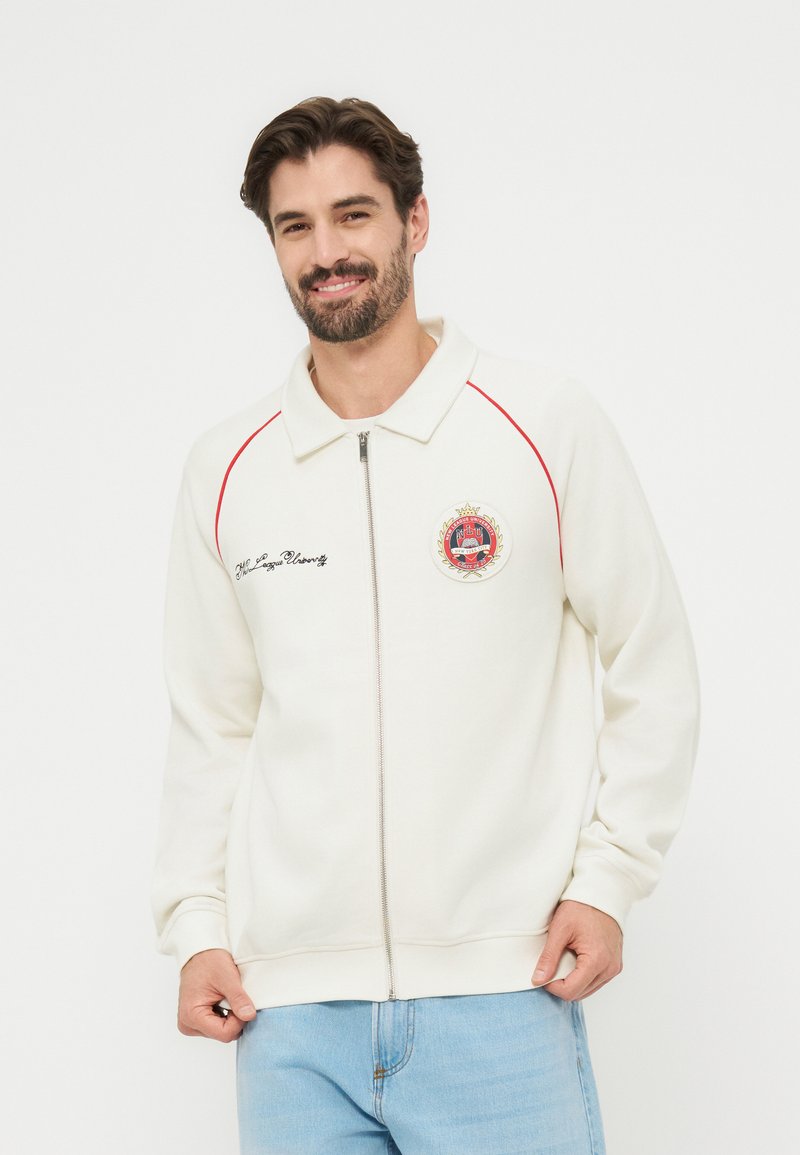 Άνδρας που χαμογελά φορώντας λευκό μπουφάν με φερμουάρ, κόκκινη διακόσμηση, κεντημένο έμβλημα και ανοιχτόχρωμα τζιν, μπροστά σε απλό φόντο.