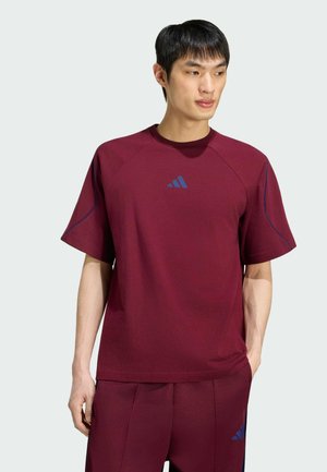 Hombre que lleva una camisa de manga corta y pantalones burdeos a juego con logotipos de Adidas en azul oscuro, de pie con las manos en los bolsillos, mirando hacia la derecha.