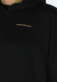 Svart hoodie i mjukt tyg med en böjd guld "SWEDEMOUNT"-logotyp på bröstet. Designen har en rymlig huva och en lös passform.