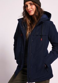 Veste imperméable bleu marine avec capuche, fermetures éclair et à pression, et deux poches latérales ; comprend une poche zippée sur la poitrine gauche et des accents rouges.
