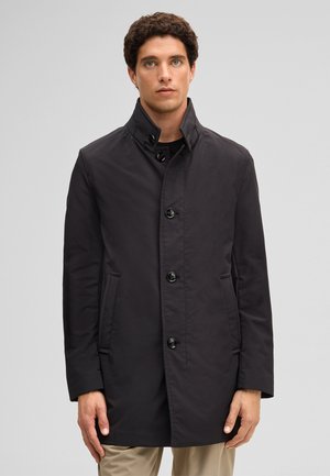 Man draagt een zwarte jas met knopen en een opstaande kraag, gecombineerd met beige broek, staand tegen een effen grijze achtergrond.