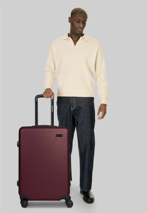 Man in crèmekleurige trui en donkere spijkerbroek, staand, met de handgreep van een grote kastanjebruine koffer met vier wielen op een grijze achtergrond vasthoudend.