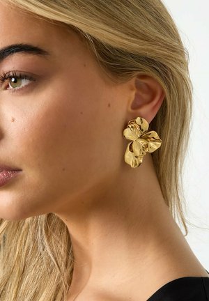 Frau mit blondem Haar, die an ihrem linken Ohr einen großen, glänzenden, goldfarbenen Ohrring in Form einer Blume vor einem einfarbigen Hintergrund trägt.