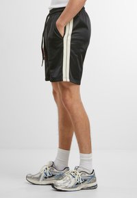 Shorts de sport noirs en tissu lisse, avec deux rayures verticales crème de chaque côté, dotés de poches latérales et d'une taille élastique.