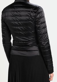 Veste noire matelassée avec des coutures horizontales, portée par une personne vue de dos, associée à une jupe plissée noire.