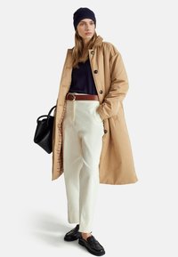 Béžový dlouhý kabát, námořnický top, bílé kalhoty, hnědý opasek, černé mokasíny, černá kabelka. Hladká látka a knoflíky na kabátu, viditelná károvaná podšívka.
