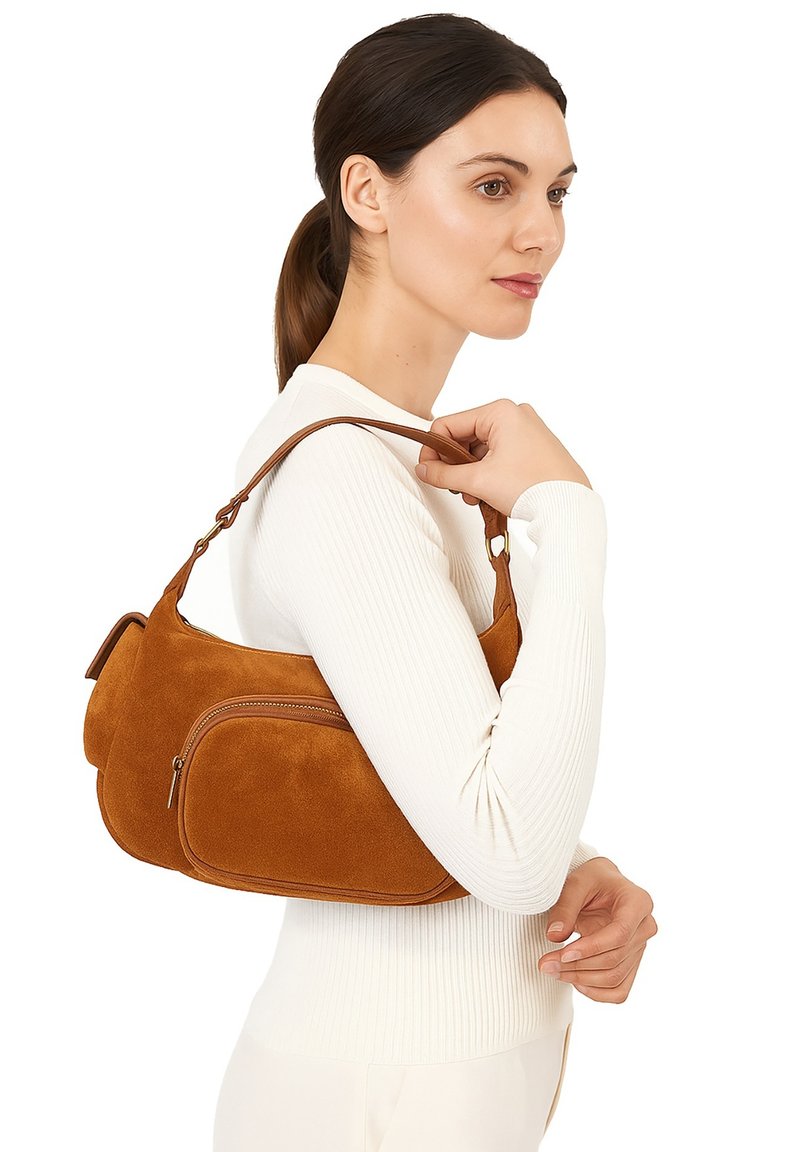 Bruine suede handtas met een gebogen vorm, twee ritssluitingen en een lange schouderriem, vastgehouden door een persoon in een witte geribbelde top.